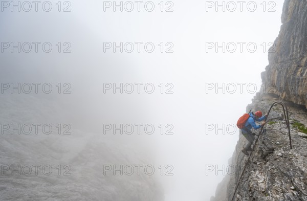 Mountaineer climbs a ladder on the Via Ferrata Oliva Detassis via ferrata in fog, cloudy steep rock faces, Brenta Mountains, Trentino, Italy