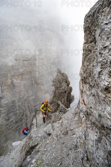 Mountaineers climb the Via Ferrata Oliva Detassis via ferrata in fog, steep mountains covered in clouds, Brenta Mountains, Trentino, Italy