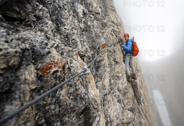 Mountaineers on a steep rock face on the Via Ferrata Oliva Detassis via ferrata in fog, steep mountains covered in clouds, Brenta Mountains, Trentino, Italy
