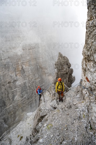 Two mountaineers climb the Via Ferrata Oliva Detassis via ferrata in fog, steep mountains covered in clouds, Brenta Mountains, Trentino, Italy