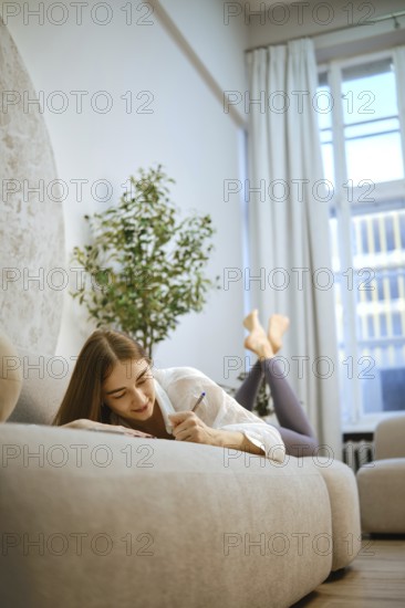 A young woman is lying on a sofa, writing in a notebook with a smile. The room has large windows letting in natural light. A plant is placed nearby, adding to the indoor space