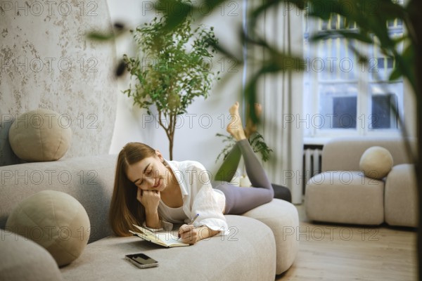 A young woman lies on her stomach on a couch in a modern living room. She is writing in a notebook with a pen while relaxing. The room has natural light coming through the window