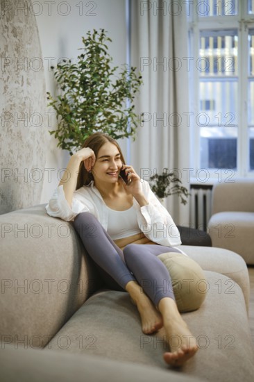 Barefoot woman is seated on a couch in a room with soft lighting. She is smiling while talking on the phone