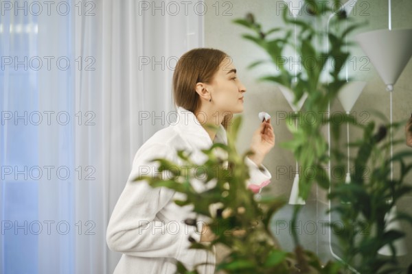 Cute woman in white bathrobe stands in a bathroom with cotton sponge in hand surrounded by green plants. Soft light filters through the curtains, creating a fresh atmosphere