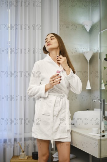 Woman stands in a bathroom wearing a robe. She holds a bottle of fragrance and is applying it to her neck. Soft light streams through curtains, creating a bright atmosphere in the room