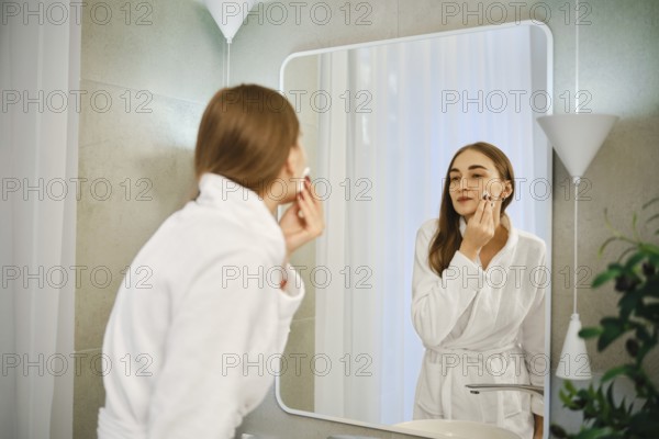 A woman stands in front of a wall mirror in a bathroom. She is washing off makeup from her face with a cotton pad