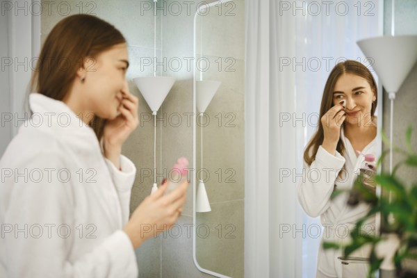 A young woman stands in front of a mirror in the bathroom. She is wearing a bathrobe and removes makeup from her face using micellar water