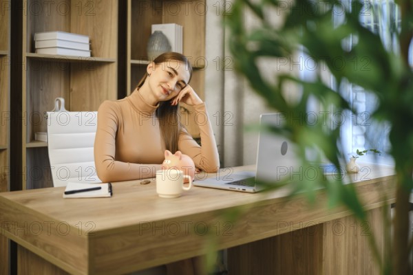 Dreamy woman sitting at a wooden desk in an office. She rests her head on her hand while looking at a laptop. A white mug and a piggy bank are placed nearby on the desk