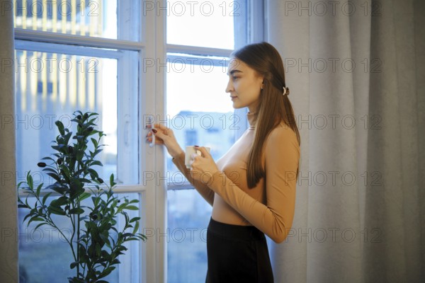 Cute woman stands by a window in a city apartment holding a coffee cup. She looks outside as the evening light fills the room. A plant is nearby, adding a touch of greenery