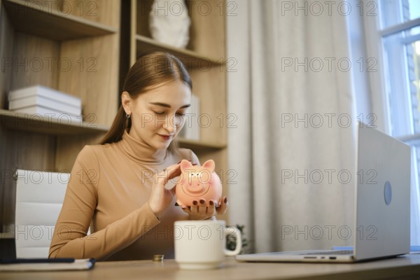 A woman sits at a desk in a home office and smiles while looking at a piggy bank. She is engaging in saving money, with a cup and laptop nearby. Sunlight is coming through the window
