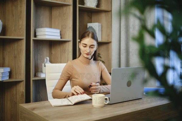 A woman is seated at a wooden desk with a laptop open in front of her. She is writing notes in a notebook. The office has shelves with boxes and a potted plant nearby