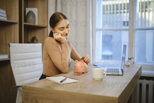 A woman is sitting at her desk in an office. She looks thoughtful while holding her chin in one hand. A piggy bank and a laptop are in front of her. A notebook and a cup are also on the desk