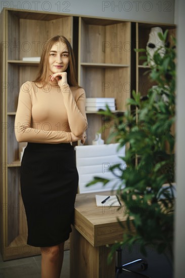 A woman stands in an office with wooden shelves behind her. She has long hair and wears a fitted top with a skirt. A green plant is in the foreground. The setting has a relaxed and inviting vibe