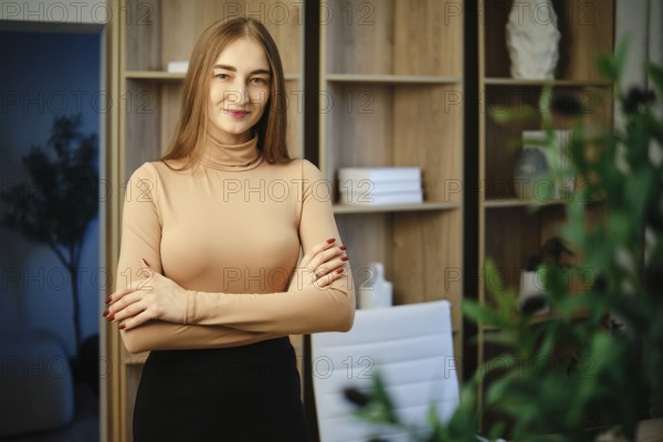 A woman with long hair stands in a modern room with wooden shelves. She has her arms crossed and has a calm expression. The room features plants and shelves with decor items