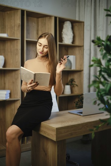 A woman sits on a wooden desk in an office. She is writing in a notebook while holding a pen. A laptop is open on the desk. Books and plants are visible on the shelves