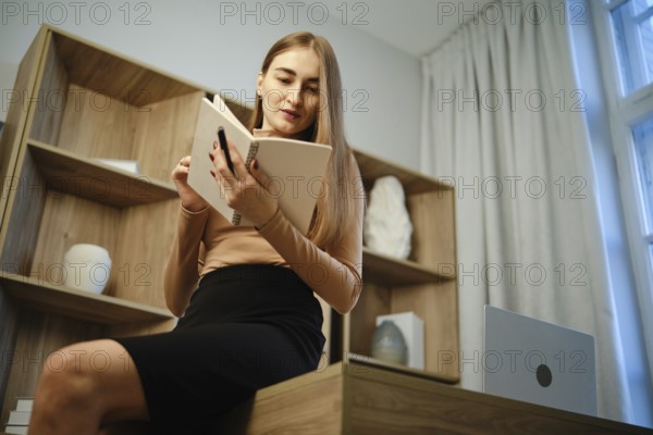 A woman sits on a wooden desk with a notebook in hand. She is focused on reading while surrounded by a modern interior. A laptop sits nearby, and natural light comes through the windows