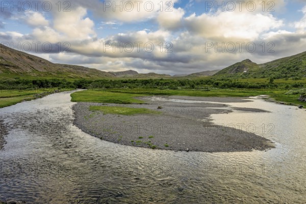 A calm river snakes through a green landscape under a cloudy sky with mountains in the background in the estuary area on the northern Varanger Peninsula, Berlevåg, Finnmark, Norway