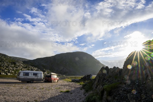 A car with roof box with old Eriba Touring Troll caravan at sunset next to rocks and a mountain in the background, Berlevåg, Finnmark, Norway