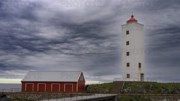 White lighthouse next to a red wooden building against a cloudy sky at the sea Kjølnes Fyr, small windows and railings in a rural area, Berlevåg, Finnmark, Norway