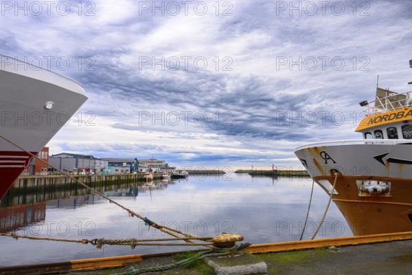 View of quiet harbor with various boats and buildings, two fishing trawler fishing vessels anchor in the harbor, calm water reflects the cloudy sky Berlevåg, Finnmark, Norway