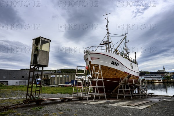A large boat fishing trawler fishing vessel on a dry dock is positioned against a cloudy sky in the harbor, Berlevåg, Finnmark, Norway