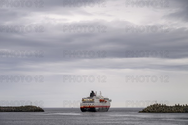 The Hurtigrouten Hurtigruten ship Kong Harald sails from Berlevag harbour, on the horizon with a cloudy sky, Berlevåg, Finnmark, Norway