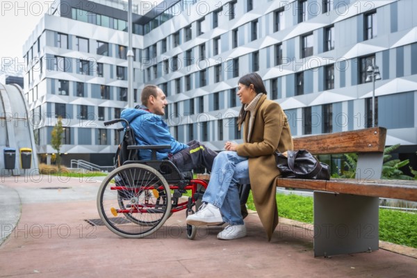 Young man living with a disability sitting in a wheelchair and having a joyful conversation with a woman sitting on a public park bench in an urban setting