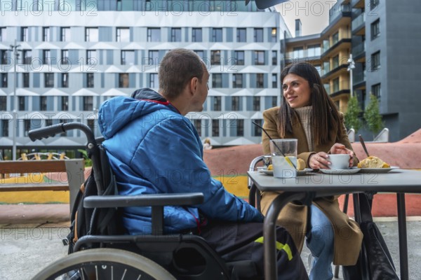Man with disability in wheelchair talking with female friend while having coffee and pastry at an outdoor urban cafe focusing on inclusion and support