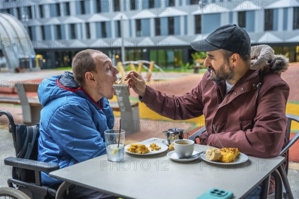 Man in wheelchair being fed by caregiver at an outdoor cafe table, showing compassion, inclusion and everyday support in an urban park setting during a casual meal