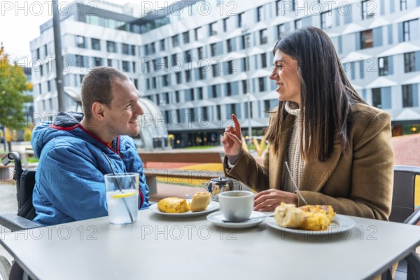 Man with a disability in a wheelchair discussing and smiling with his female social worker over drinks and snacks at an outdoor cafe, representing care, support, and inclusion