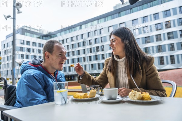 Woman feeding a smiling man with a disability, enjoying a meal and coffee at an outdoor cafe, embodying moments of care, companionship, and shared enjoyment in an urban setting