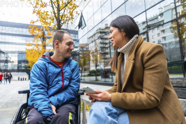 Man in wheelchair smiling and attentively listening to a woman sitting beside him on a bench, having a friendly and inclusive conversation in an urban area with autumn trees