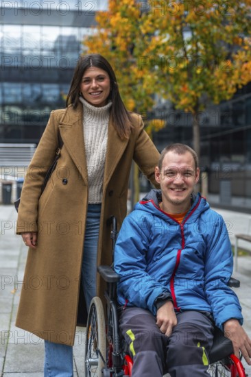 Young woman pushes her smiling partner in a wheelchair along an accessible urban pathway amid autumn foliage, sharing support, independence and joyful outdoor moments