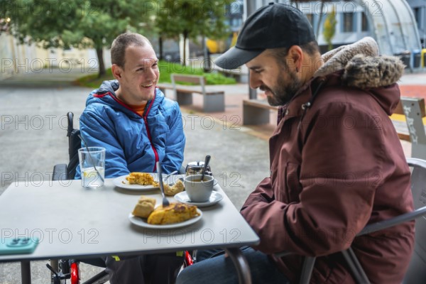 Young man with a disability smiling and relaxing in a wheelchair at an outdoor cafe table, enjoying a coffee and conversation with a friend, showing lifestyle, inclusion, and companionship