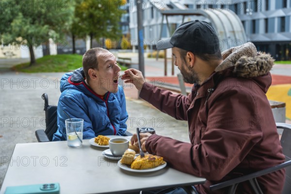 Man tenderly feeding a young boy in a wheelchair at an outdoor cafe, representing care, support, and inclusion for people with disabilities in public spaces