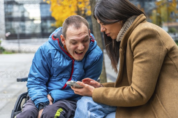 Disabled man in a wheelchair smiling with a female caretaker enjoying a fun moment together while looking at a smartphone screen outside during autumn