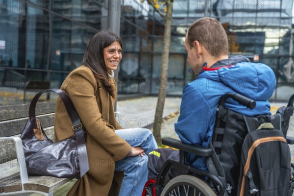 Woman sitting on bench chatting and laughing with a boy in a wheelchair in a city park, warm inclusive moment of support, friendship, empathy and everyday urban life