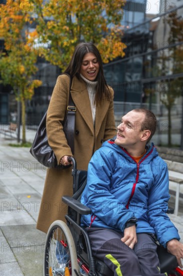 Cheerful caregiver standing behind a young man in a wheelchair, pushing him along an urban sidewalk during a colorful autumn day, smiling and talking together