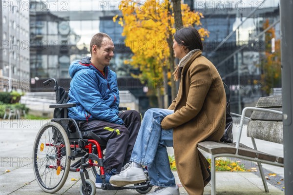 Young man living with disability in a wheelchair smiling and talking with a woman sitting on a park bench during autumn, showcasing support, inclusion, and friendship in an urban setting