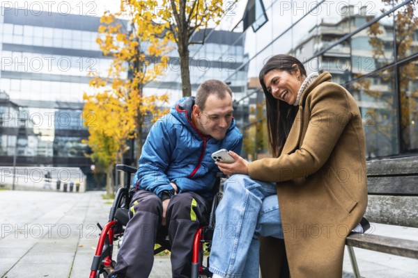 Man in a wheelchair and woman sitting on a bench, sharing a laugh and looking at a mobile phone screen outdoors in an urban setting with autumn foliage