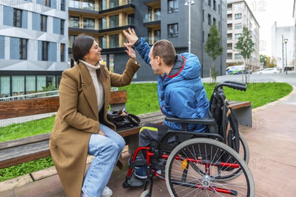 Female caregiver high fiving a young man in a wheelchair while sitting on a city park bench, sharing a joyful, supportive moment of inclusion and friendship outdoors