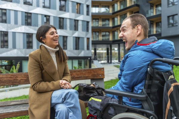 Young woman and man with a physical disability in a wheelchair sharing a joyful moment and smiling widely while communicating on a bench in front of modern buildings