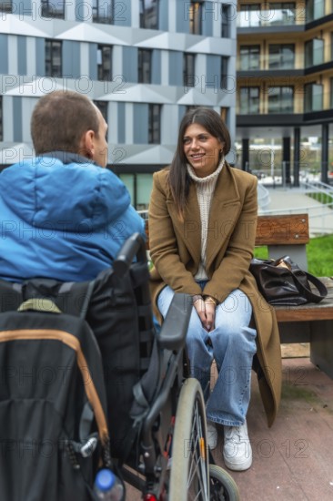 Young woman sitting on a park bench talking and smiling with a man in a wheelchair, showing inclusion, friendship and support in an urban outdoor setting during daytime