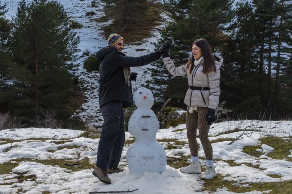Young couple enjoying winter vacation, high fiving, and smiling while building a snowman on a snowy hillside with pine trees and mountains in the background