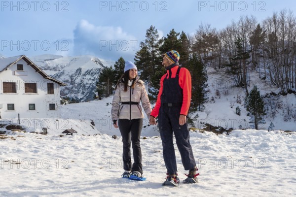 Happy couple holding hands while snowshoeing on a sunny winter day, enjoying active leisure time in a snowy mountain landscape with a rustic house and evergreen trees