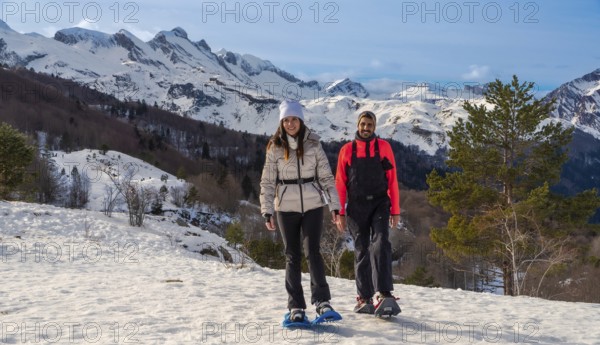 Couple snowshoeing on a snowy mountain trail, smiling and looking at the camera, with majestic snow capped peaks and clear blue sky in the background, embodying outdoor recreation and winter travel