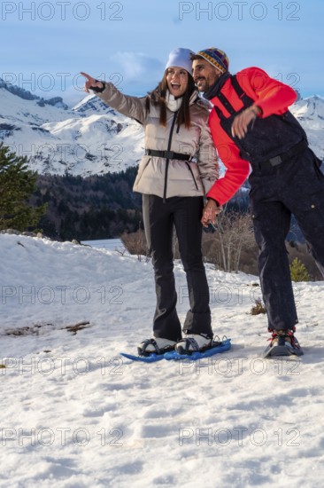 Smiling couple snowshoeing across snowy mountain slopes, pointing toward a breathtaking winter panorama under blue skies, enjoying active outdoor adventure and fresh alpine air