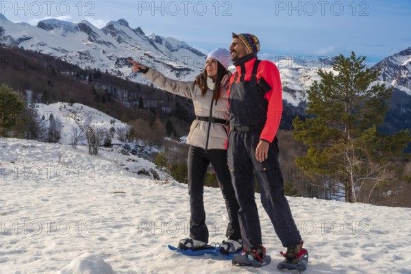 Happy couple snowshoeing in a snowy mountain landscape enjoying a winter adventure, woman pointing towards the horizon while man watches, experiencing nature and outdoor recreation together