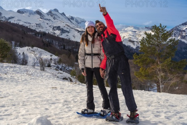 Young couple enjoying outdoor winter adventure, snowshoeing in a snowy mountain landscape under blue skies, with the man pointing excitedly towards something in the distance