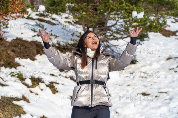 Young woman dressed in a warm jacket joyfully celebrating the outdoor winter season by throwing fresh snow into the air, creating a playful moment in a snowy forest